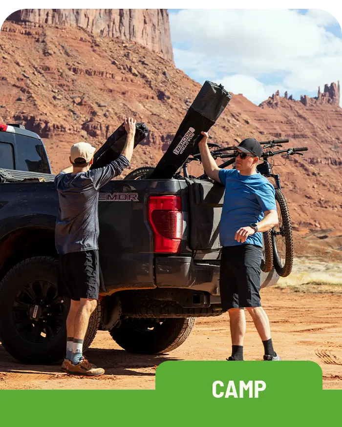 A man wearing a hat and sunglasses lifts a black Roadtrip Rocker chair from the bed of a pickup truck while another man in a blue hoodie assists. They are in a desert landscape with red rock formations in the background.