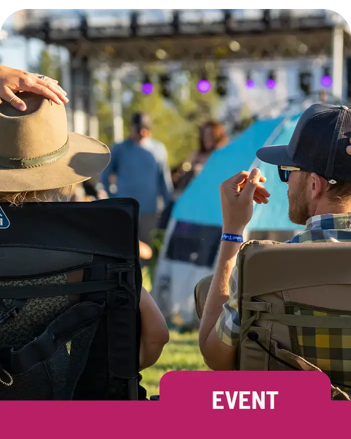 A group of people relaxes in Everywhere Chair 2 seats at an outdoor concert or festival. A man wearing a hat and a wristband leans back while another person beside him watches the stage. String lights and a large tent are visible in the background.