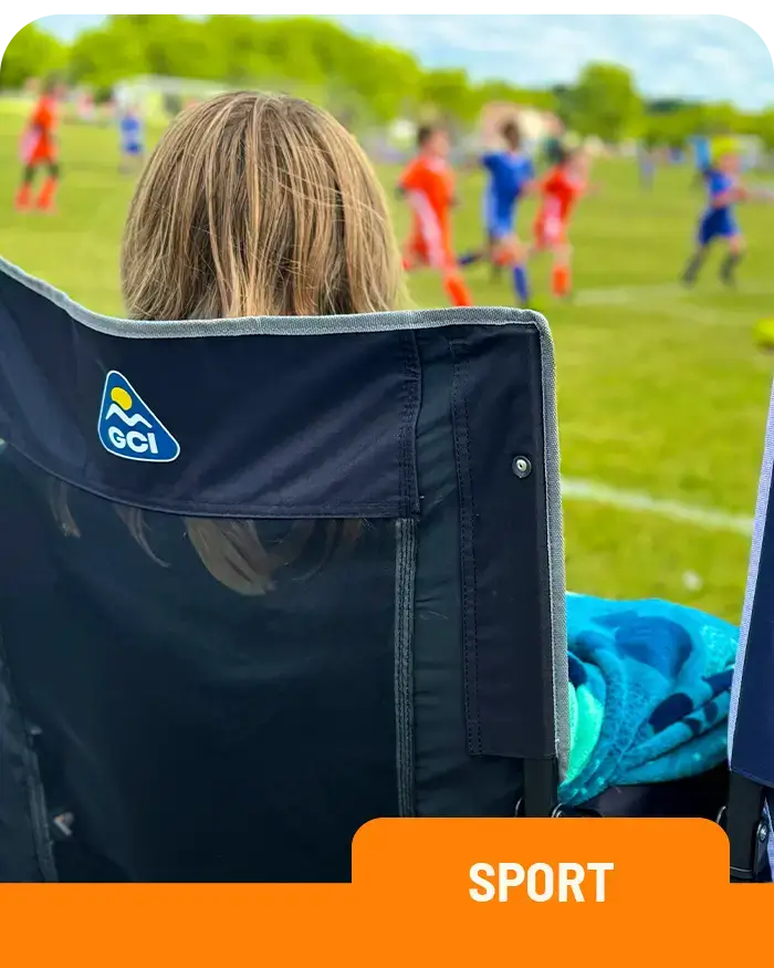 A woman with long blonde hair sits in a Stowaway Rocker chair on the sidelines of a grassy sports field, watching a youth soccer game. Players in red and blue jerseys run across the field in the background.