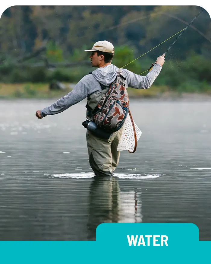 A man in a gray hoodie, fishing vest, and cap stands knee-deep in a river, fly fishing. He holds a fishing rod with one arm extended as he casts his line, with a scenic forested backdrop.