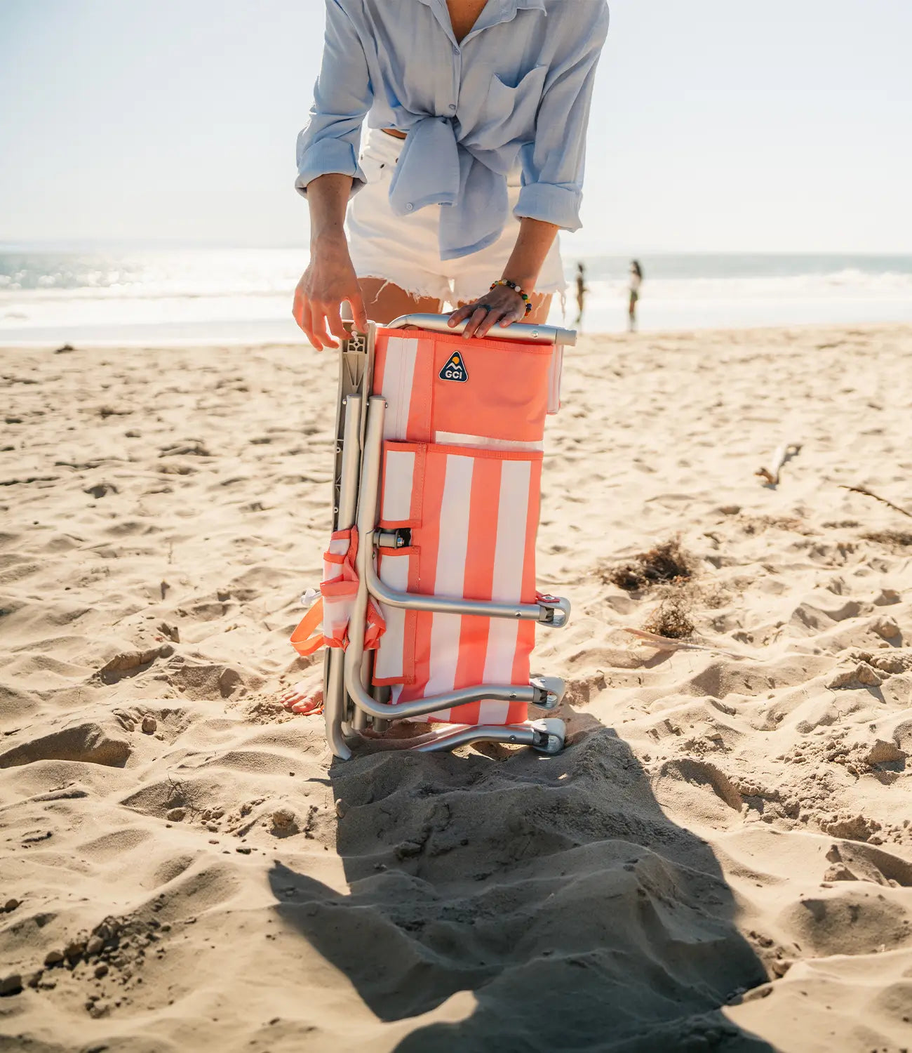 A person on a sandy beach is folding a bright orange and white striped beach chair with a silver metal frame.
