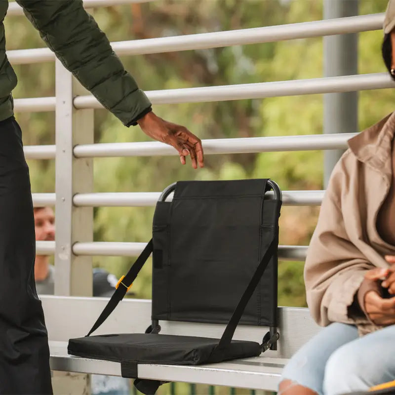 Black SitBacker stadium seat secured to bleachers, with padded backrest and straps visible as someone reaches to sit down.