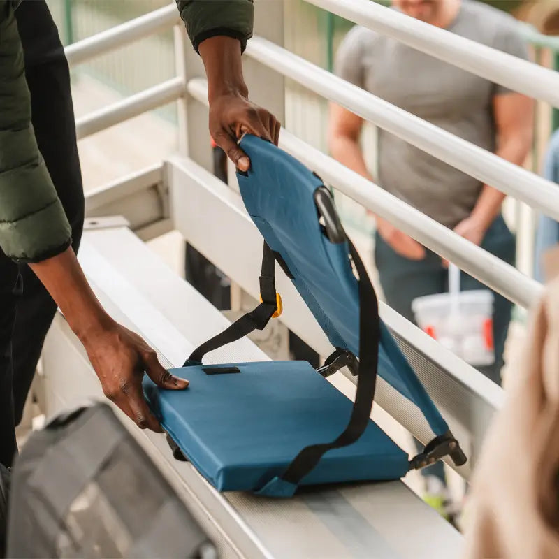 Close-up of someone unfolding a blue SitBacker seat onto metal bleachers at a sporting event.