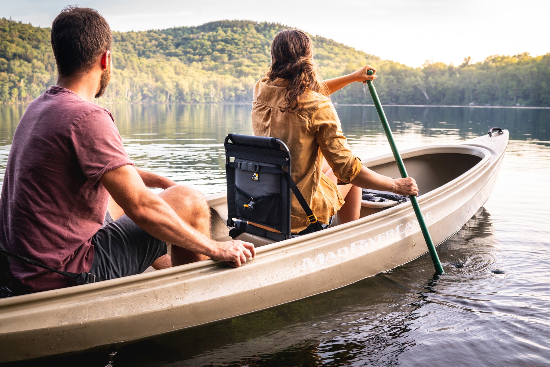 A man and woman paddle a canoe on a calm lake, with the SitBacker seat attached to the canoe for back support and comfort.