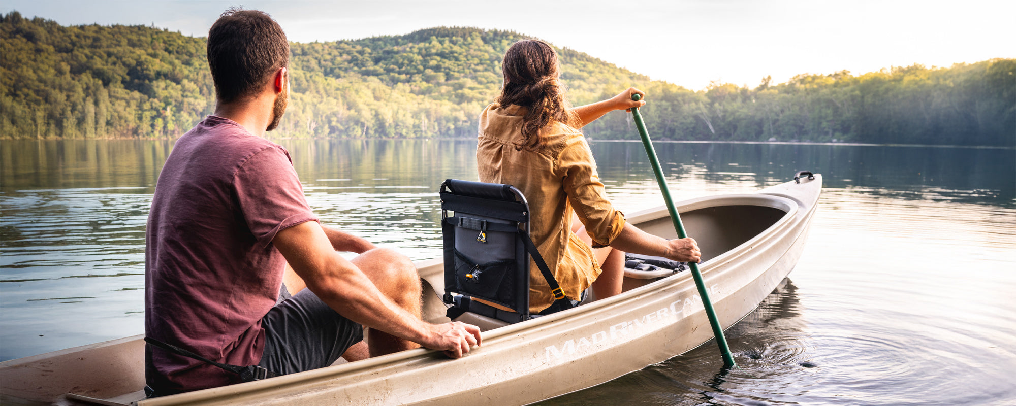 A man and woman paddle a canoe on a calm lake, with the SitBacker seat attached to the canoe for back support and comfort.