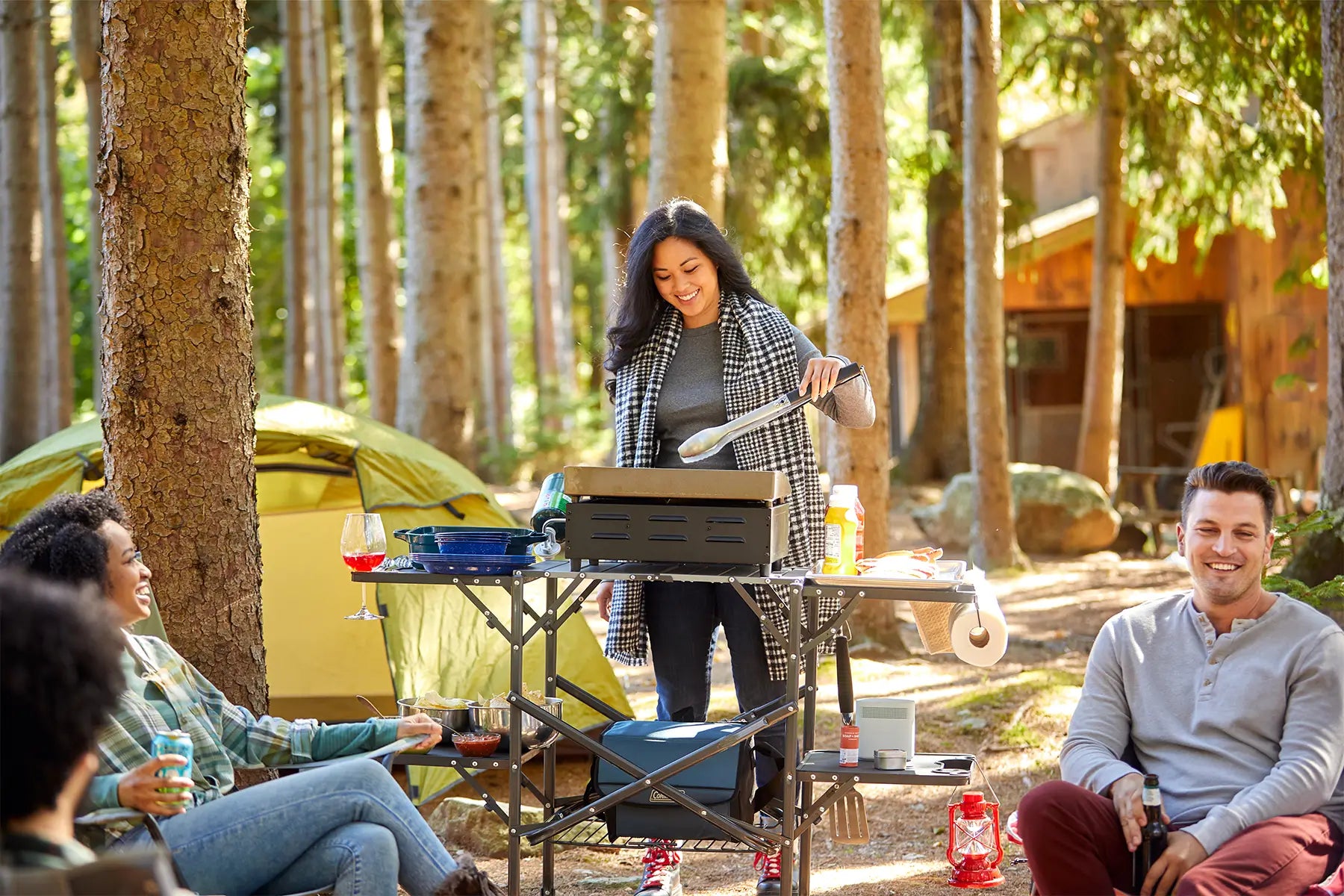 A woman grilling on the Slim-Fold Cook Station while others hang around a firepit at a campsite. 