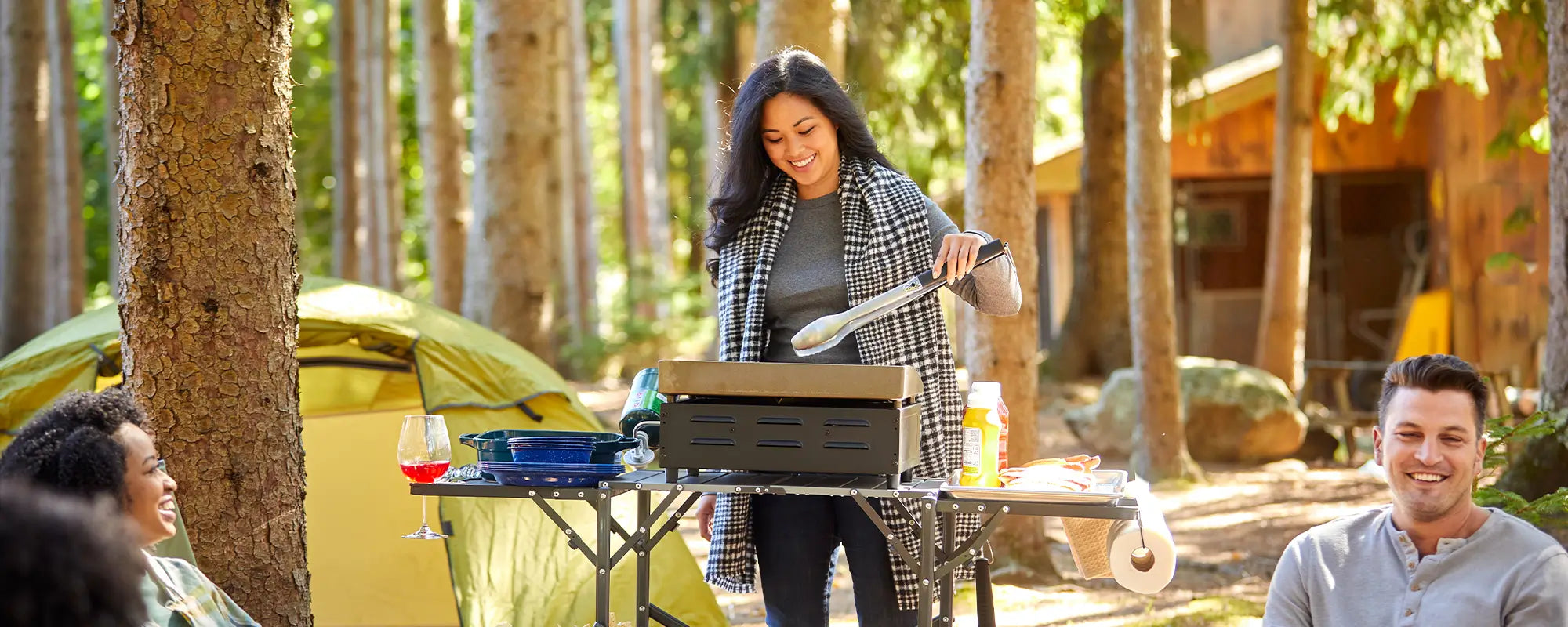 A woman grilling on the Slim-Fold Cook Station while others hang around a firepit at a campsite. 