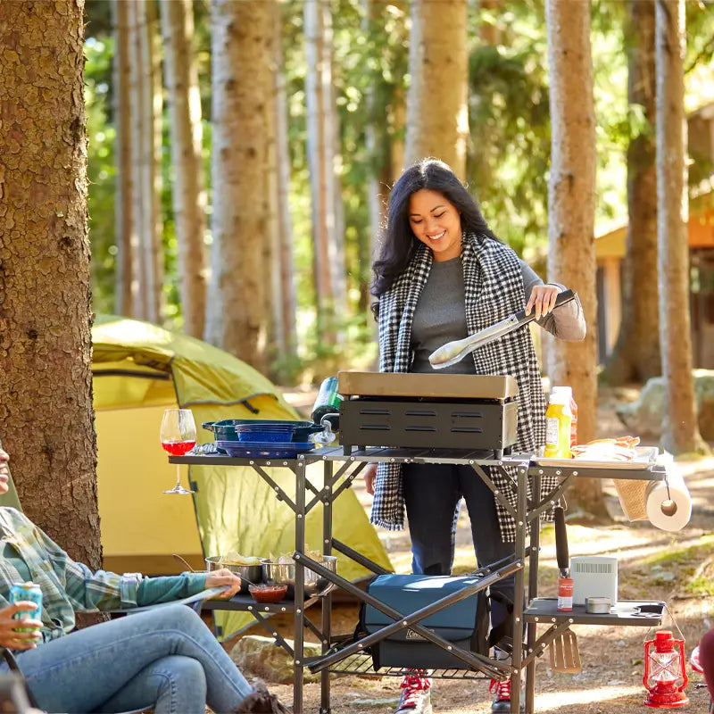 Woman grilling at campsite on slim-fold cook station surrounded by tents and trees.