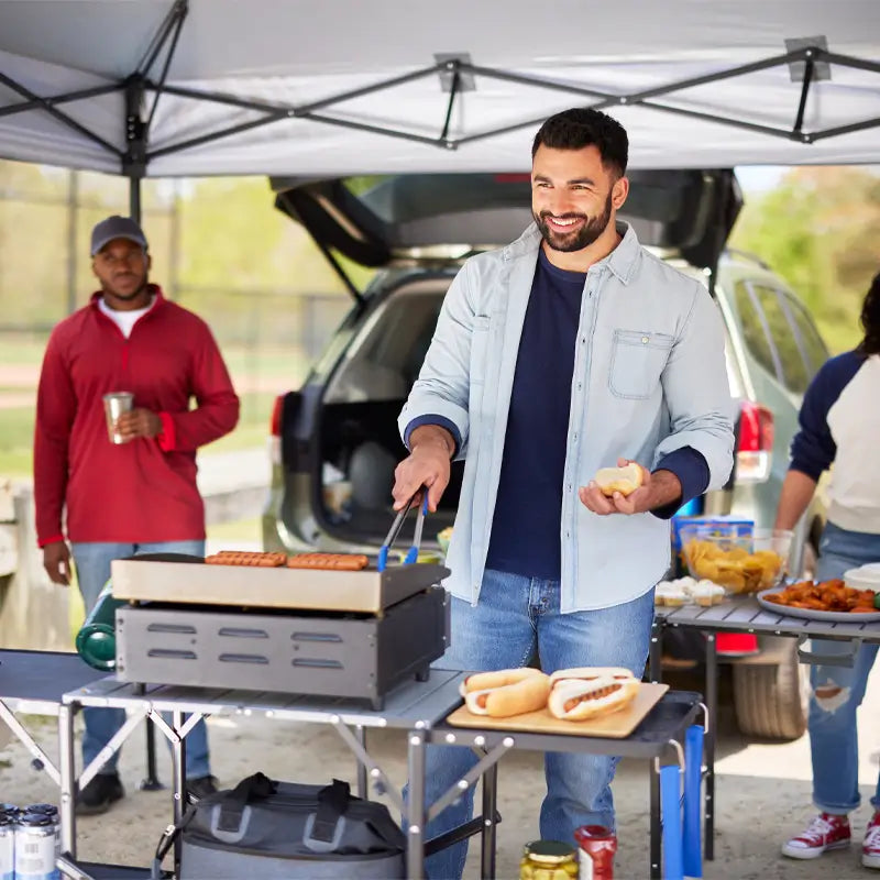 Man grilling hot dogs on slim-fold cook station during a tailgate cookout.