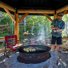 Man grilling food near a fire pit with a Slim-Fold Cook Station behind him holding buns and condiments.