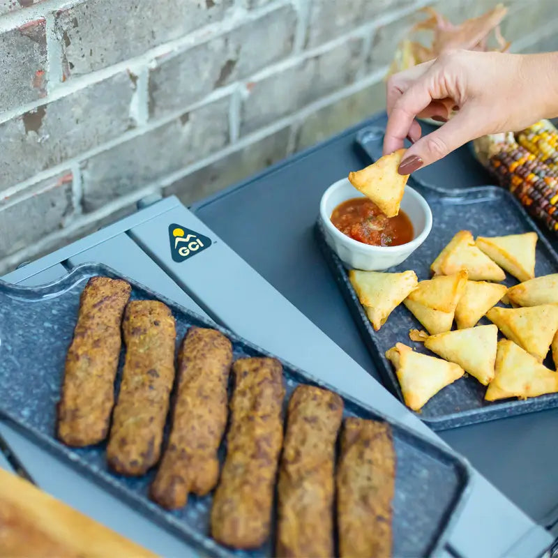 Close-up of samosas and snacks on the Slim-Fold Cook Station with a hand dipping into salsa.