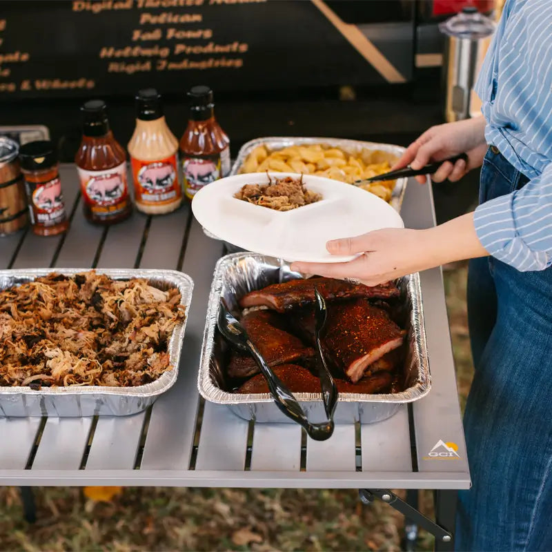 Person serving barbecue from trays on a slim-fold table at an outdoor gathering.