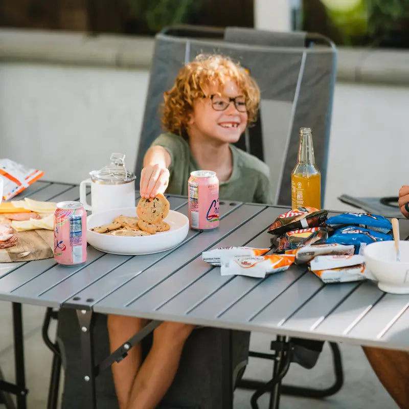 Smiling child grabbing cookies from a slim-fold table covered with snacks and drinks.