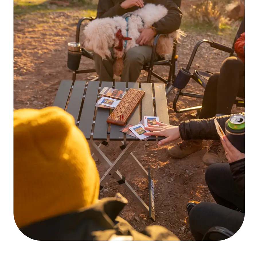 Group sitting around a Slim-Fold Table, playing cribbage and cards during a sunset camping trip.