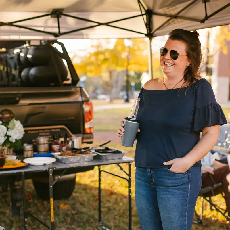 Woman smiling while holding a drink in one hand and standing beside a fully set-up Slim-Fold Table XL at a tailgate party under a canopy.