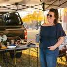 Woman smiling while holding a drink in one hand and standing beside a fully set-up Slim-Fold Table XL at a tailgate party under a canopy.