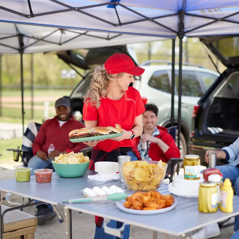 Woman in a red cap serving burgers at a Slim-Fold Table XL loaded with chips, dips, and condiments during a festive tailgate scene.