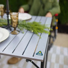 Close-up of the Slim-Fold Table XL corner showing a plated tablescape with vintage glassware and a greenery centerpiece; GCI Outdoor logo visible.