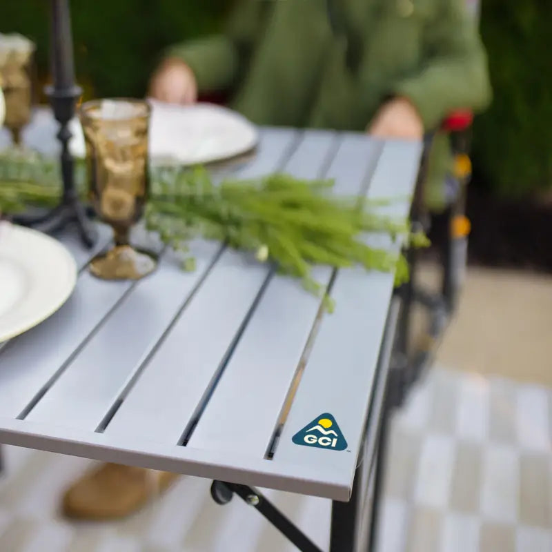 Close-up of the Slim-Fold Table XL corner showing a plated tablescape with vintage glassware and a greenery centerpiece; GCI Outdoor logo visible.