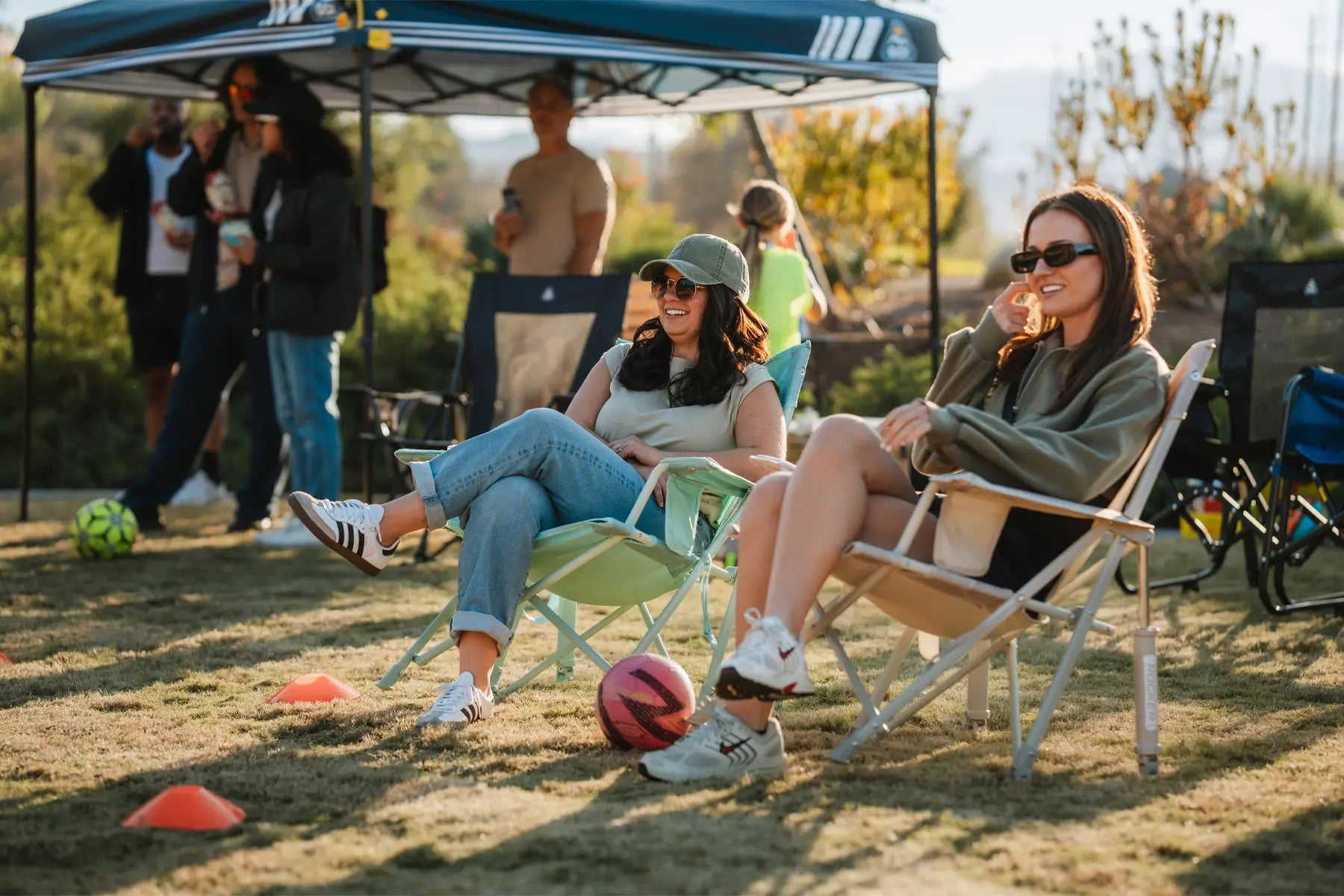 Two women sitting in comfort chairs on the sidelines, laughing with a soccer ball and cones nearby.