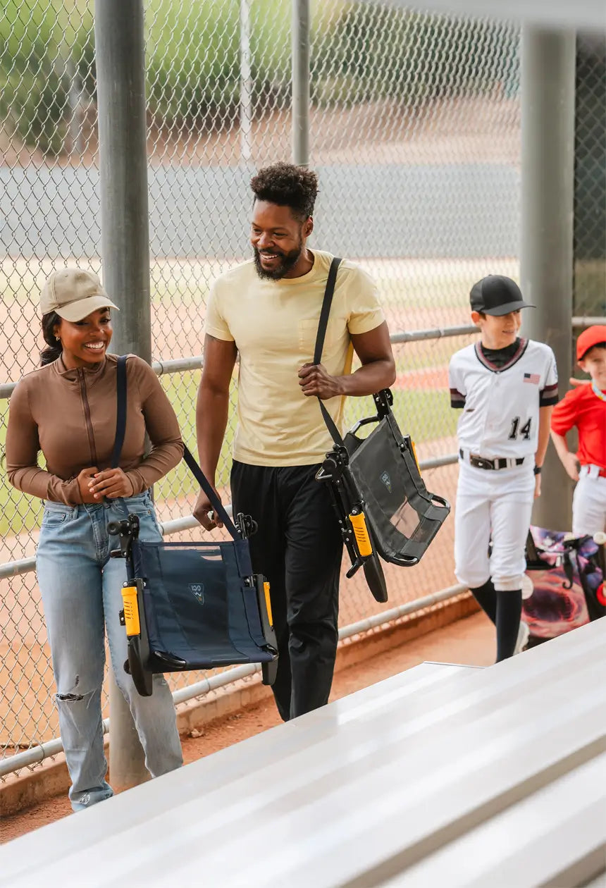 A smiling couple walks up the bleachers carrying Stadium Rock-Cliners, ready to watch a baseball game. 