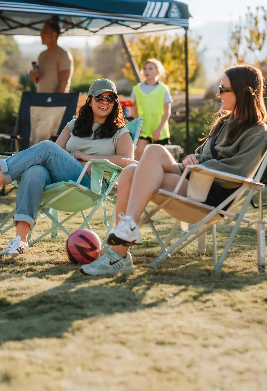 Two women relax in GCI Outdoor Comfort Pro Rockers at a youth soccer game, chatting and laughing with a soccer ball nearby under a shaded canopy.