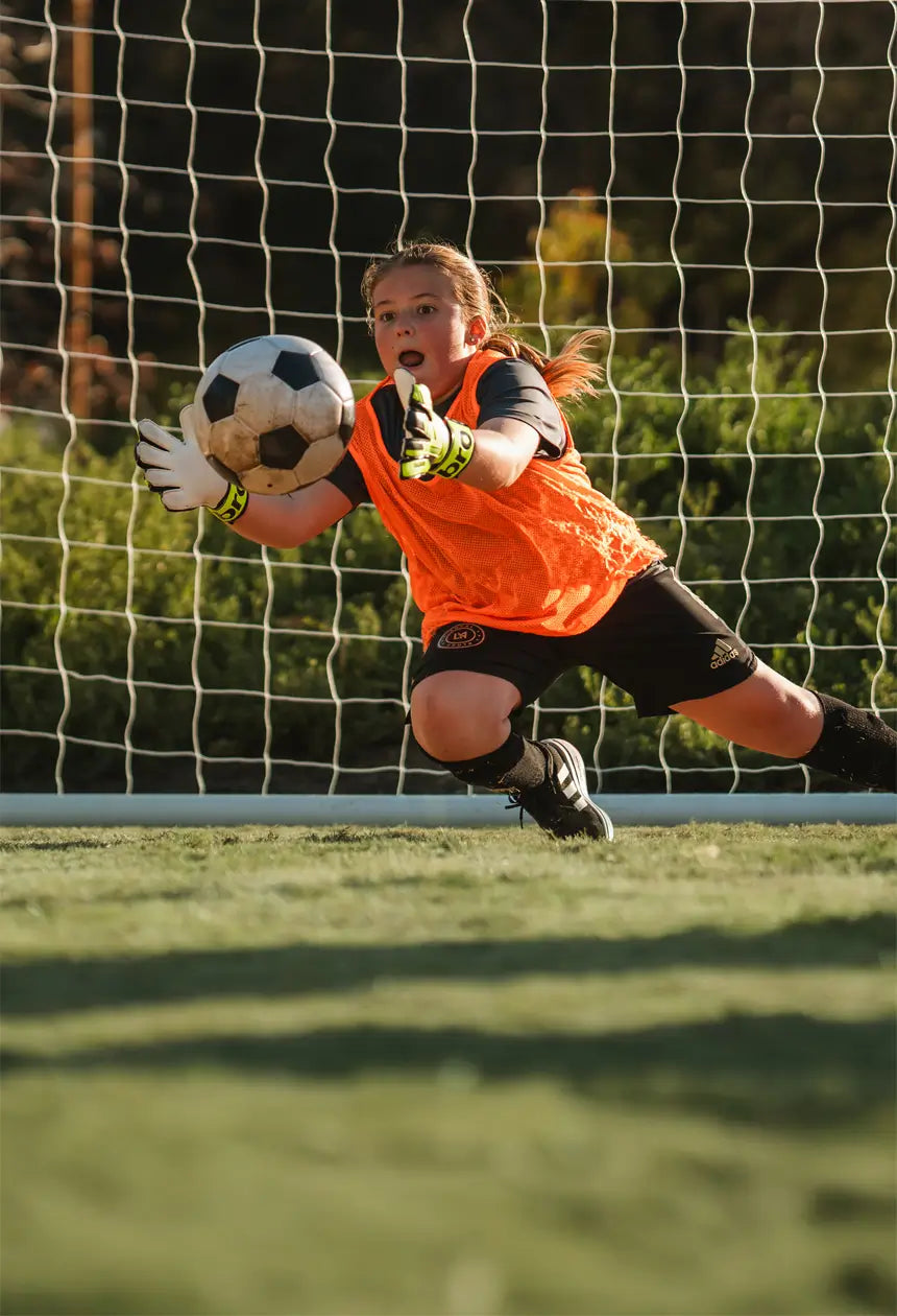  A young goalkeeper in an orange jersey and gloves dives to make a save, reaching for the soccer ball in front of the net.