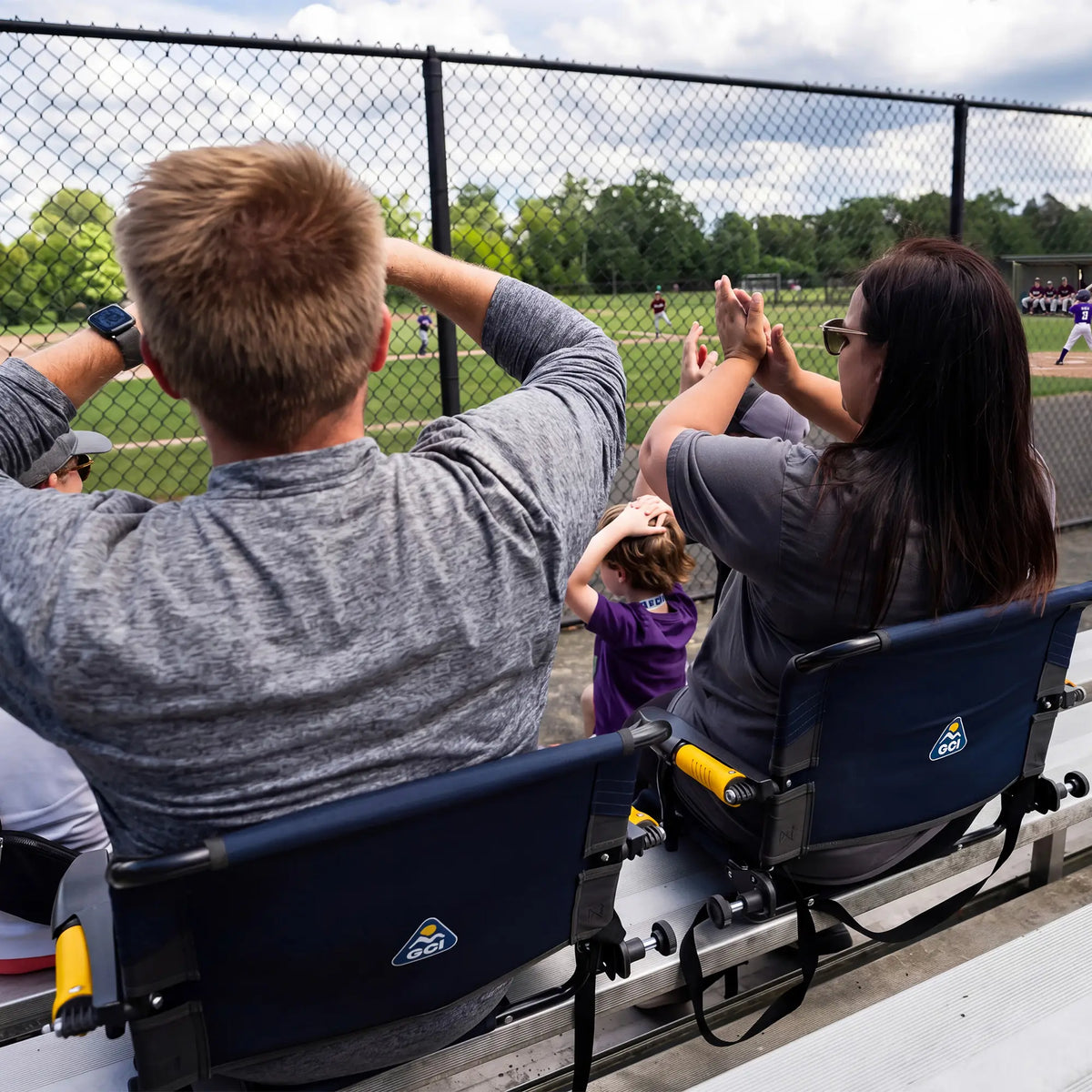 A family sits in blue Stadium Rock-cliner chairs at a baseball game, viewed from behind as they watch the action through a chain-link fence.