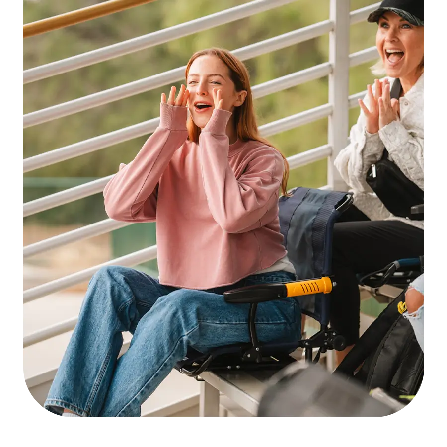 A young woman cheers while seated in a Stadium Rock-Cliner, with another excited spectator beside her. The portable chair features armrests and back support.