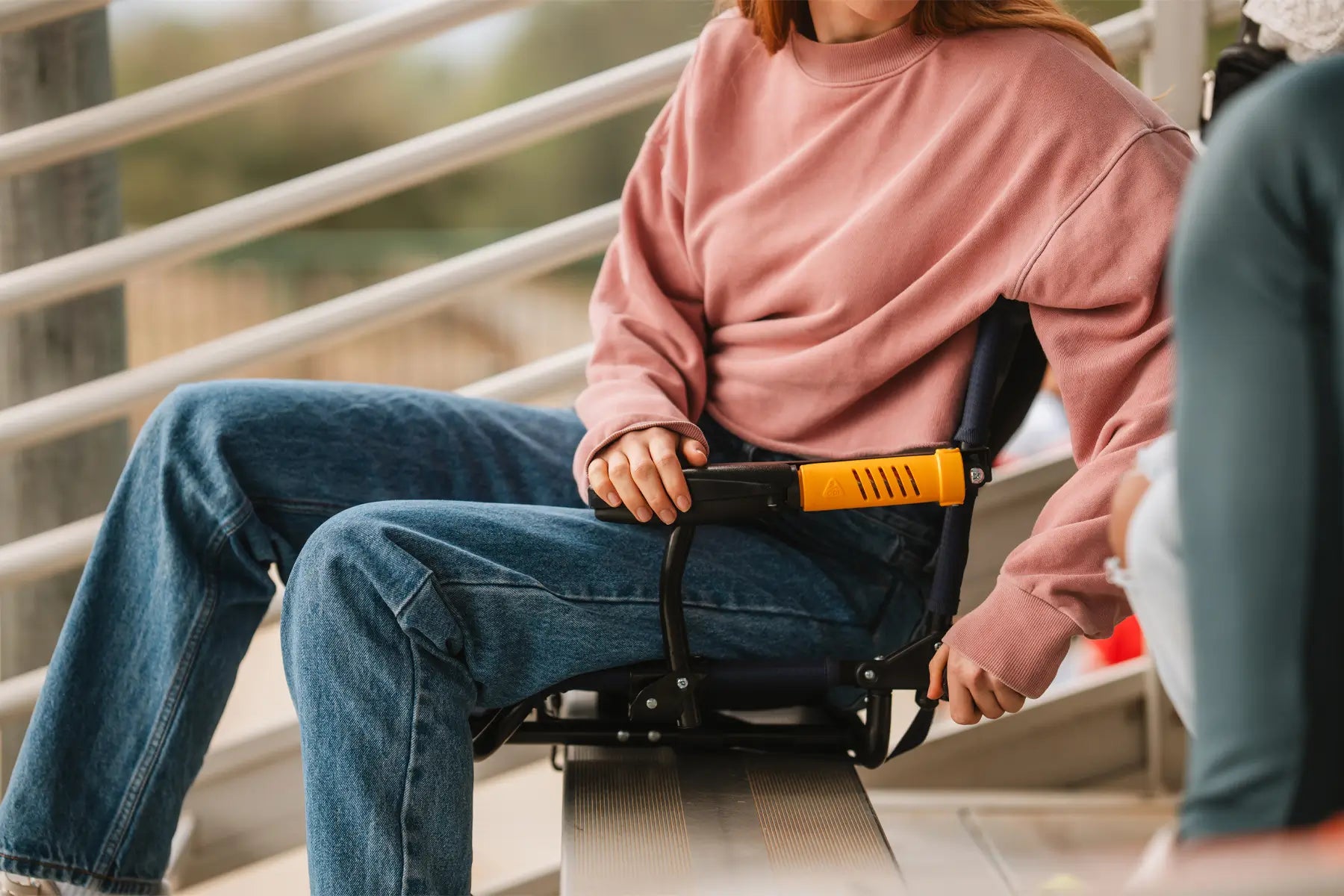 A person sits on a Stadium Rock-Cliner attached to a bleacher, adjusting the armrest for comfort while watching an event.