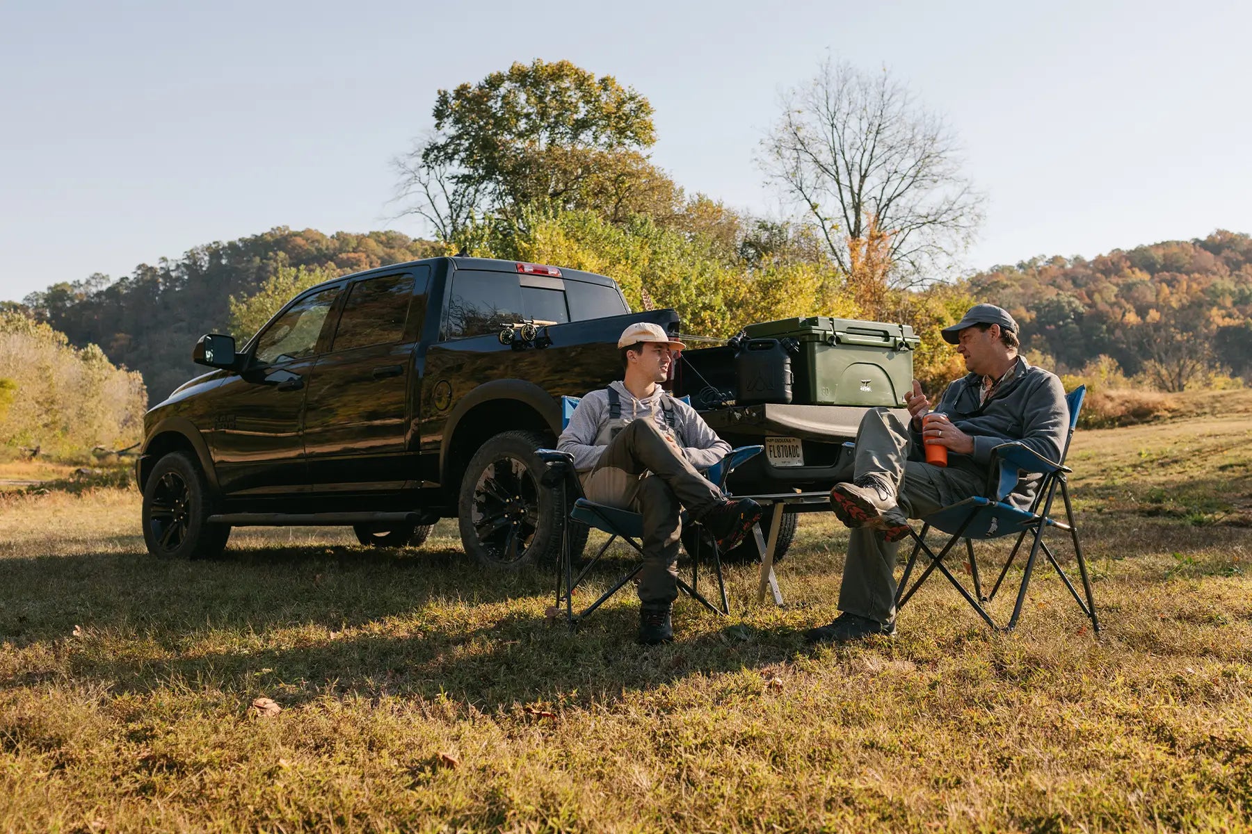 Two men sitting in gci outdoor stationary chairs in an open field next to their pickup truck. 