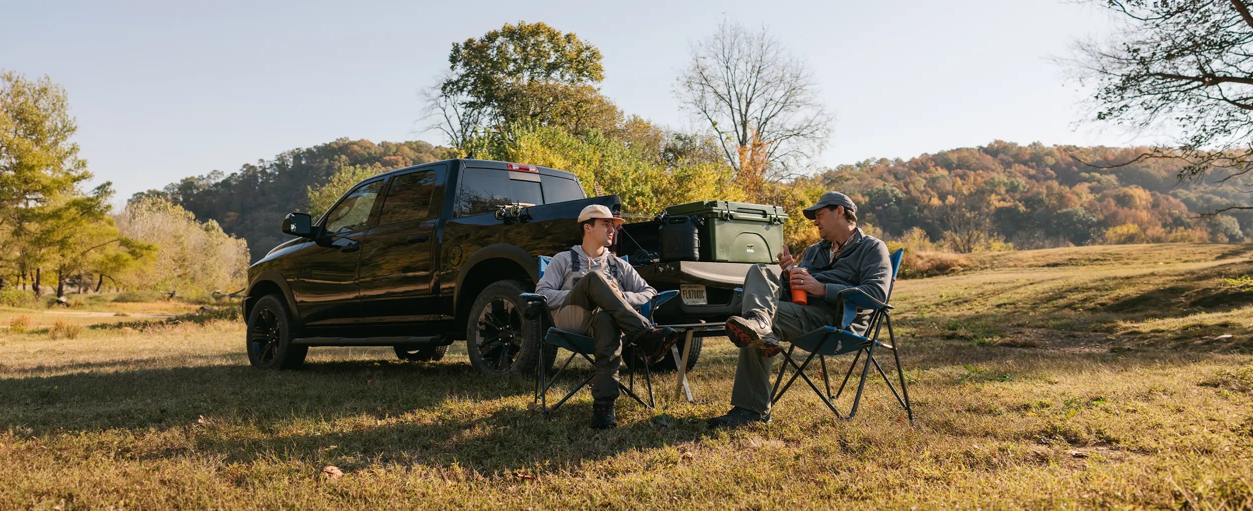 Two men sitting in gci outdoor stationary chairs in an open field next to their pickup truck. 