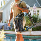 Person carrying a folded GCI Stowaway Rocker in Navy Topo with shoulder strap by a backyard pool.