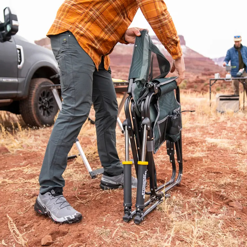 Person unfolding a GCI Stowaway Rocker in Hunter Topo on red desert terrain near a campsite.