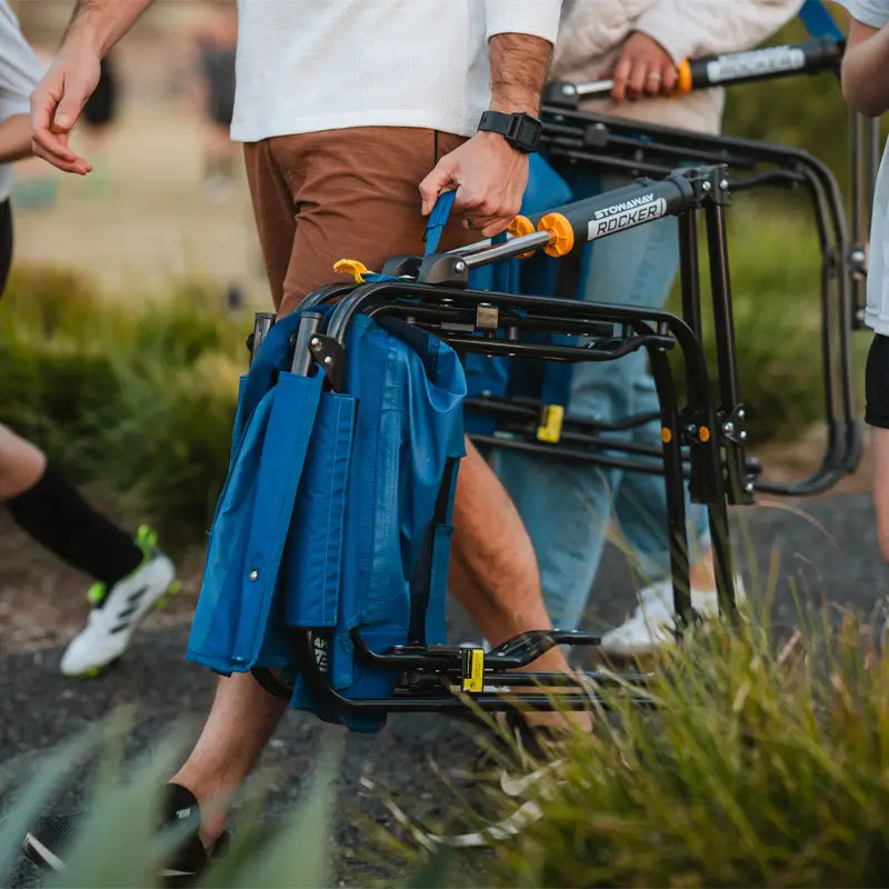 Person carrying a folded GCI Stowaway Rocker in Soft Navy, showing its compact design and easy transport handle.