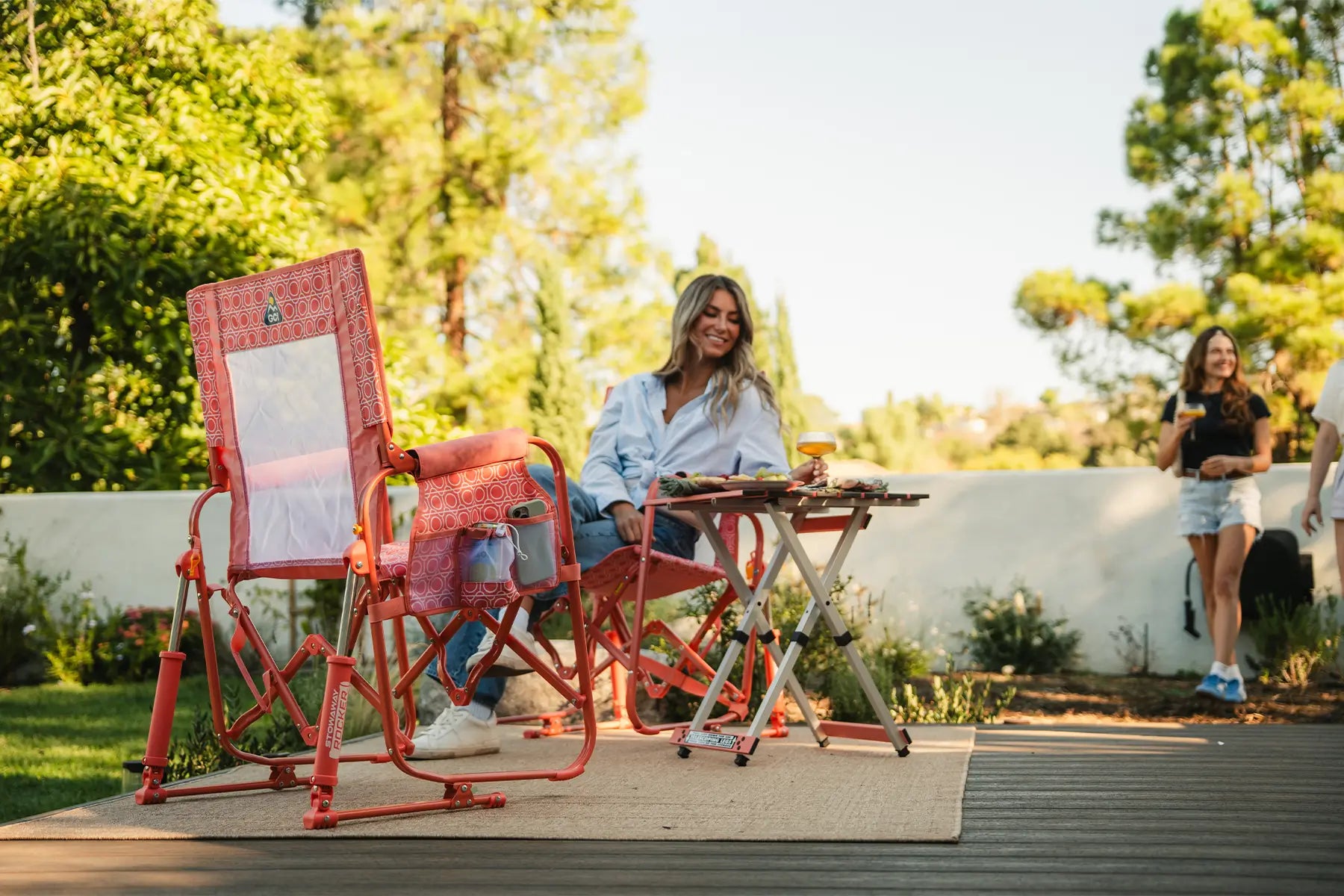 A woman sits at a small folding table on a wooden deck in a garden setting, using a pink coral Stowaway Rocker.