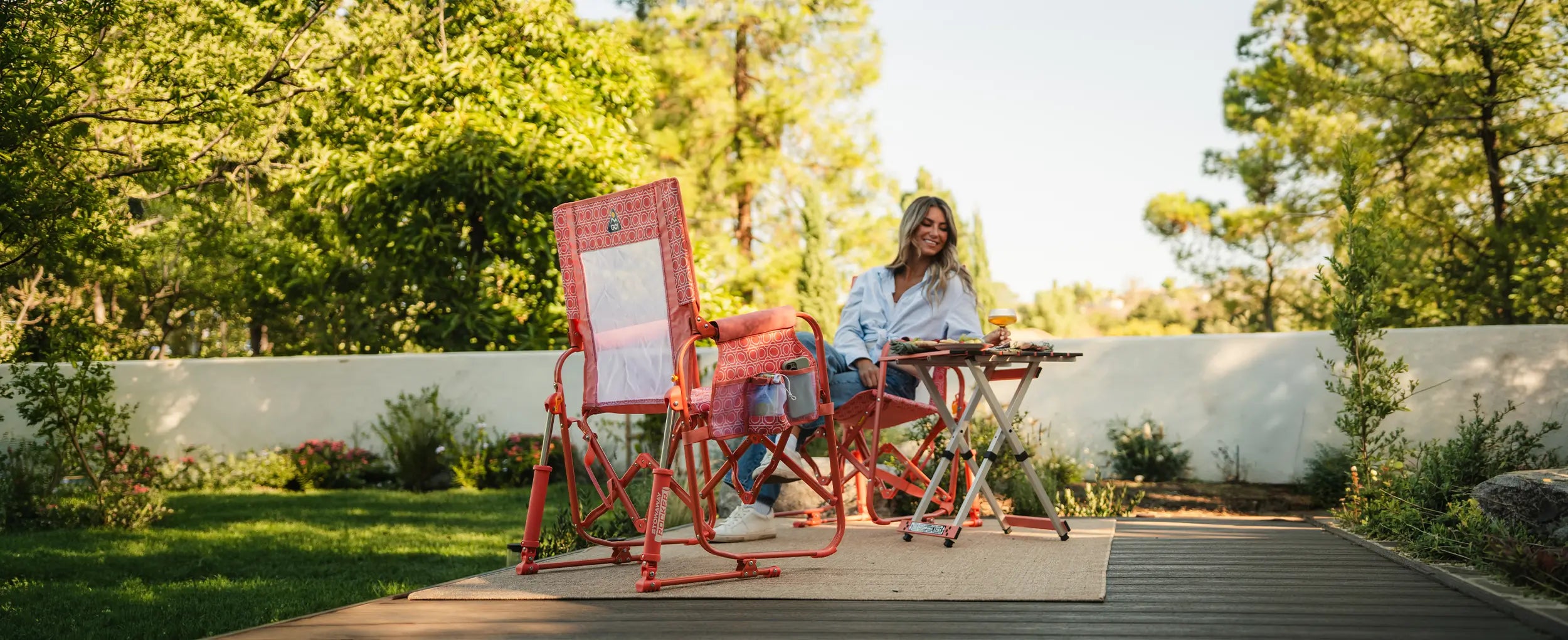 A woman sits at a small folding table on a wooden deck in a garden setting, using a pink coral Stowaway Rocker.