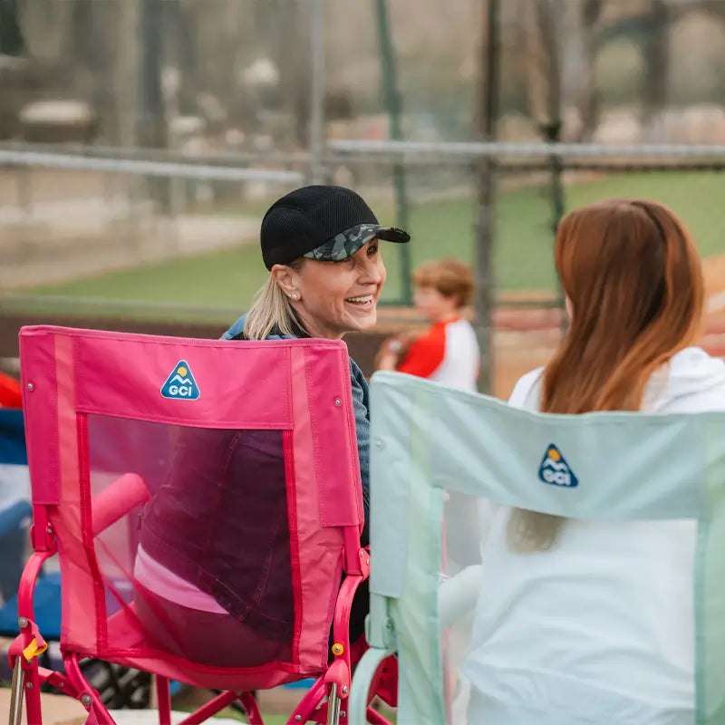 Women smile and talk while seated in pink and mint Stowaway Rockers near a baseball field.