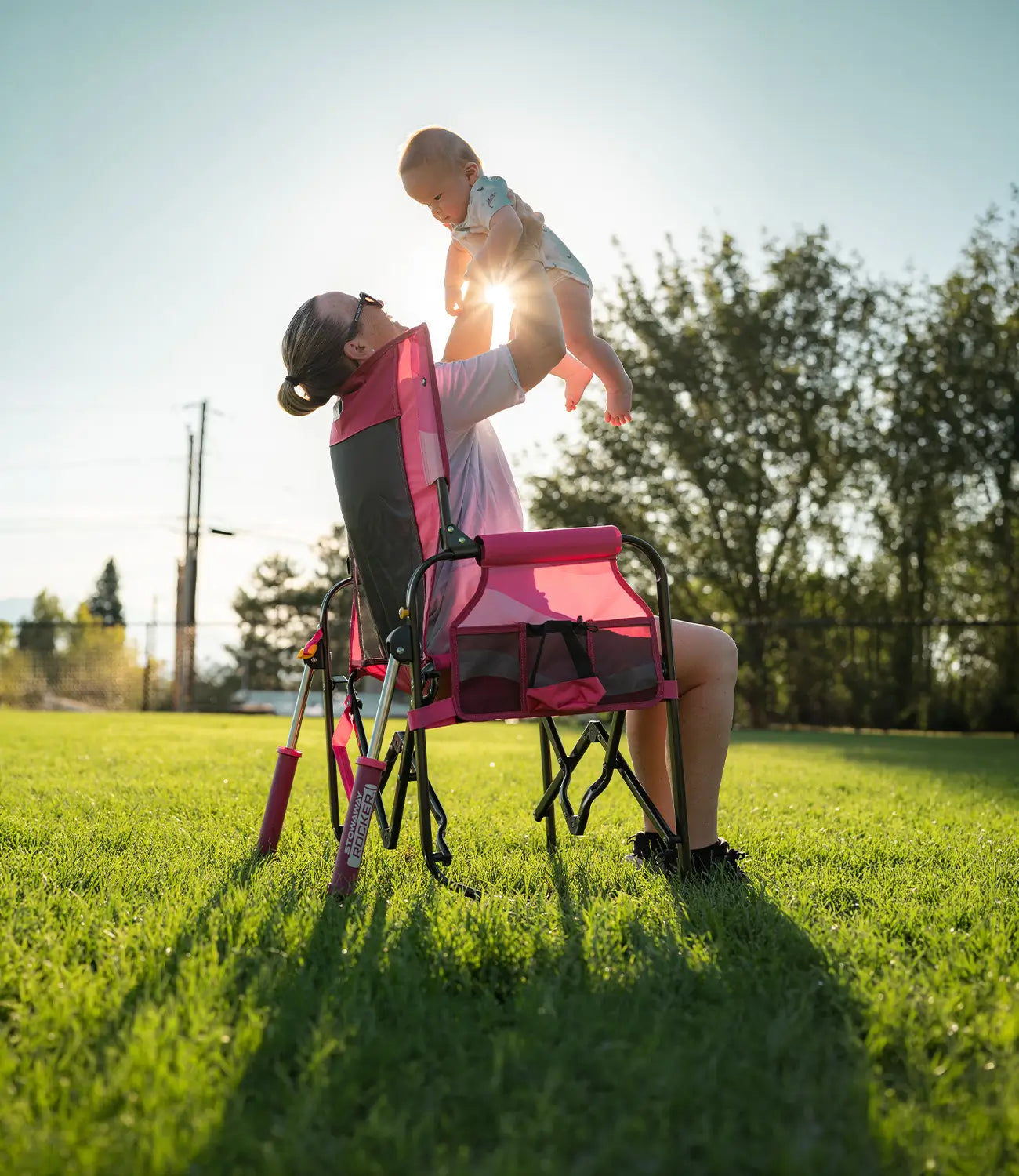 A woman sits in a pink and black Stowaway Rocker on a grassy lawn, holding a baby above her head toward the setting sun. Trees and a fence are visible in the background.