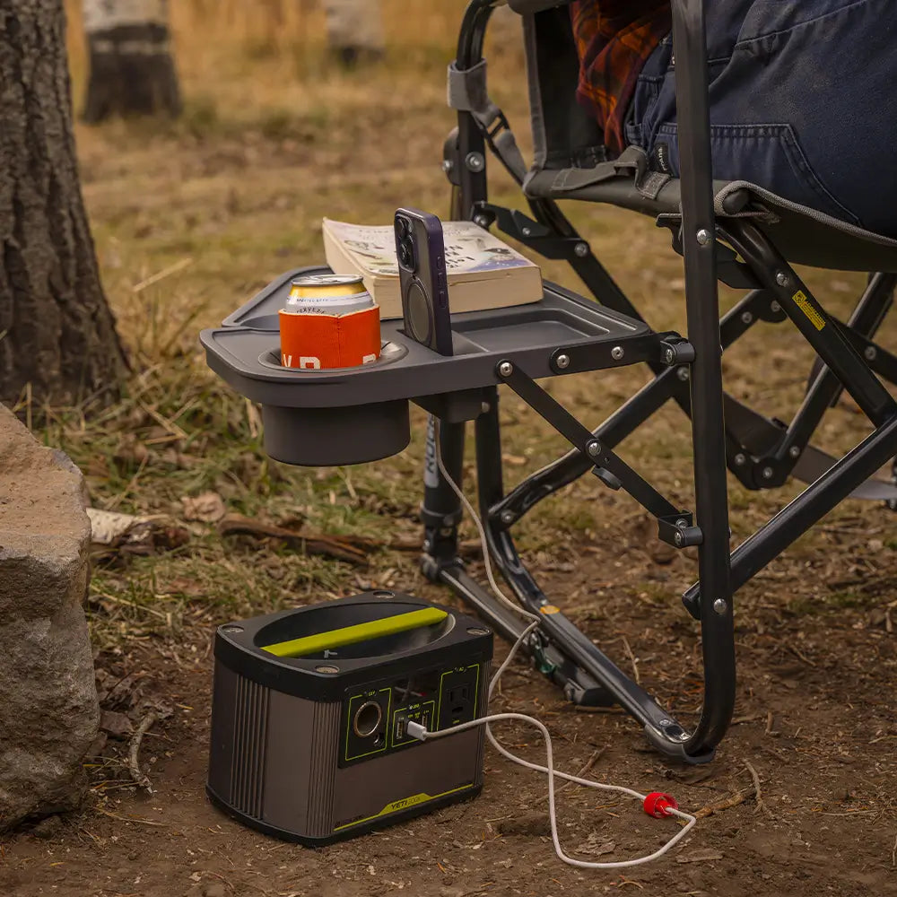Black Stowaway Rocker with Side Table holding a drink and small items beside a portable power station at a campsite.