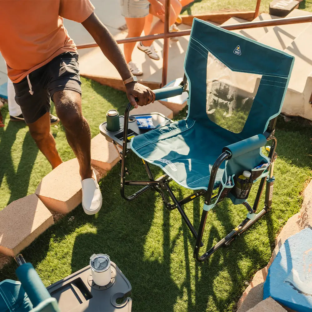 Person unfolding the Teal Stowaway Rocker with Side Table outdoors on grass near a baseball field. 