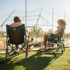 Two people relaxing in Teal Stowaway Rockers with Side Tables beside a sports field on a sunny day.