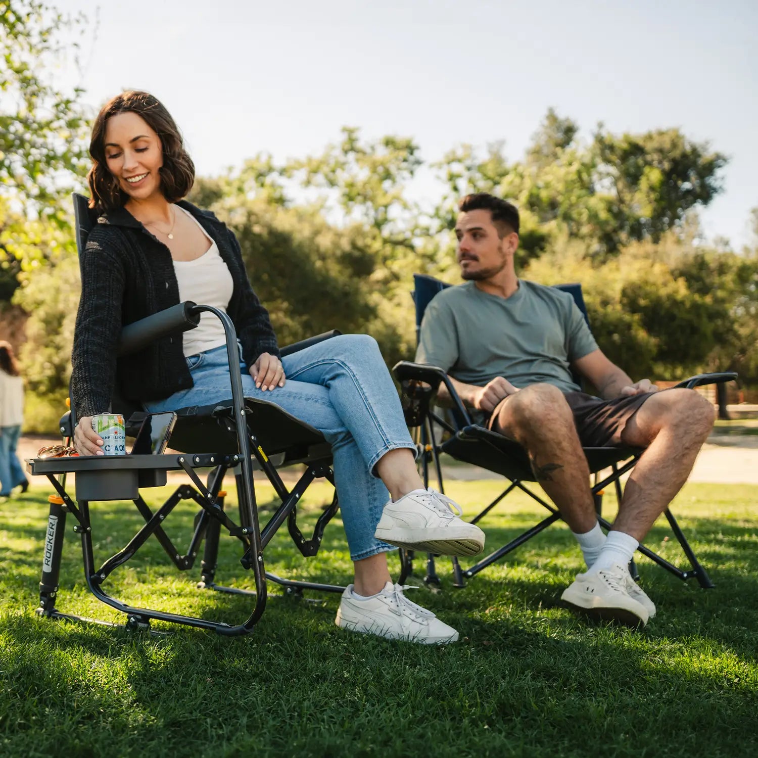 A couple sitting together at a park in their Comfort Pro Rocker and Stowaway Rocker with Side Table. 