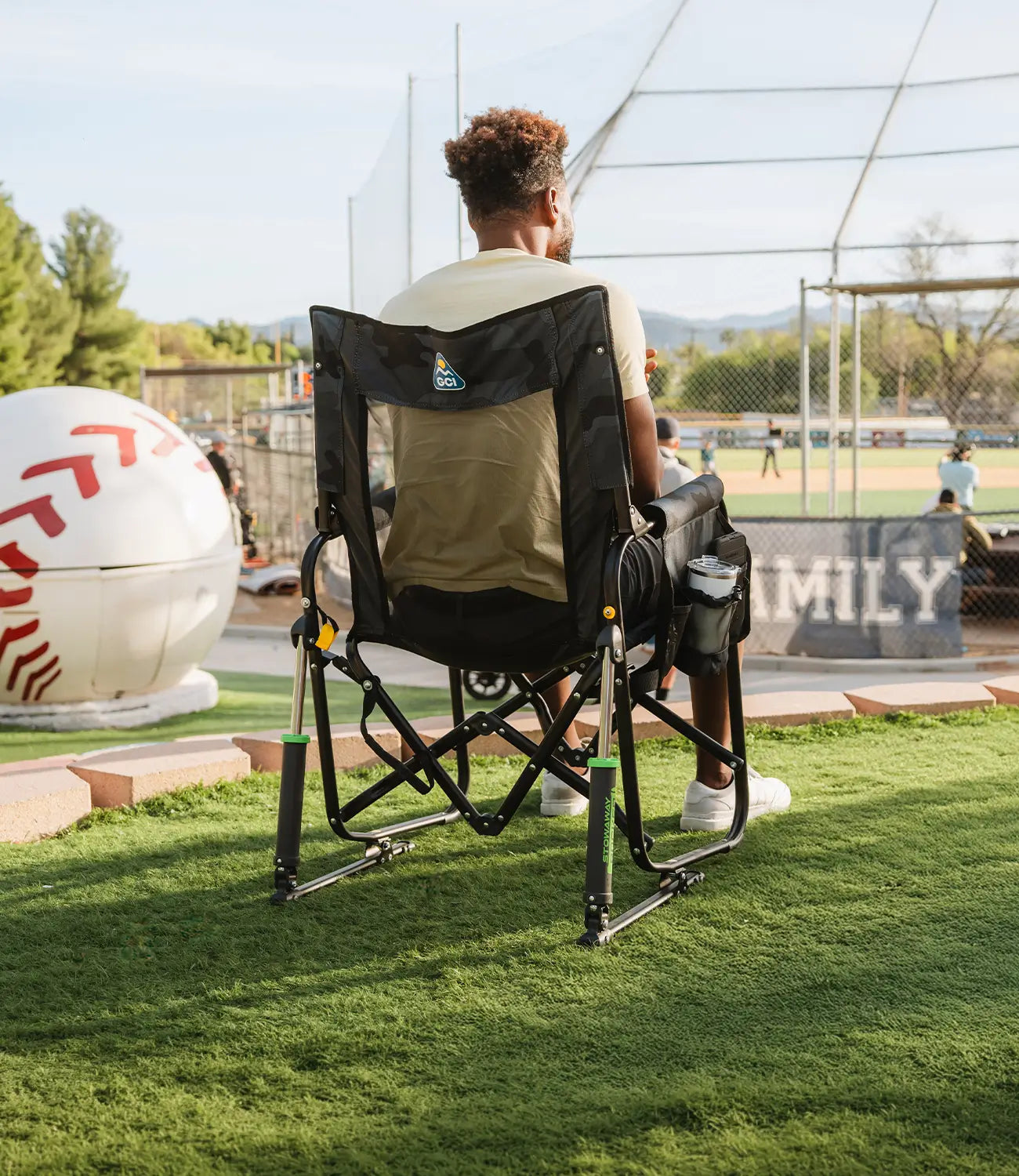 A man sitting in a stealth camo Stowaway Rocker as he watches a baseball game. 