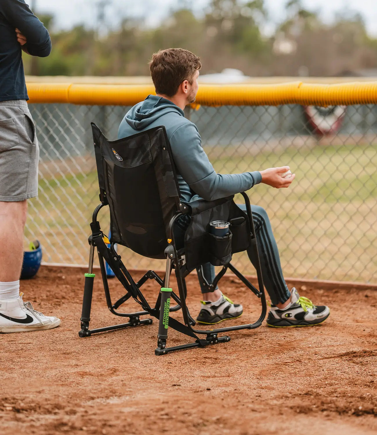 A man sitting in a stealth camo Stowaway Rocker attending a baseball game. 