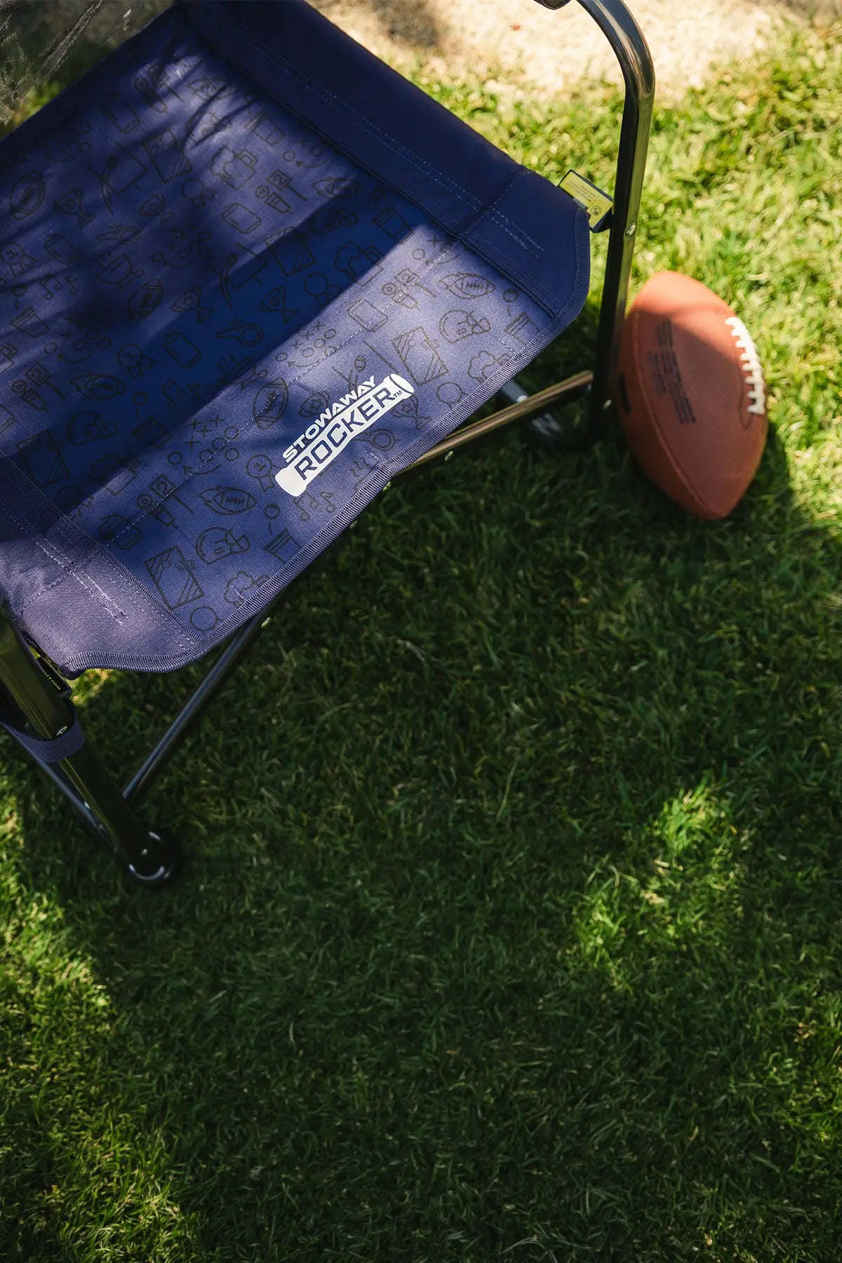 A tailgater stowaway rocker chair with a patterned seat cushion sits on green grass next to a brown football, with a tree trunk and dappled sunlight in the background.