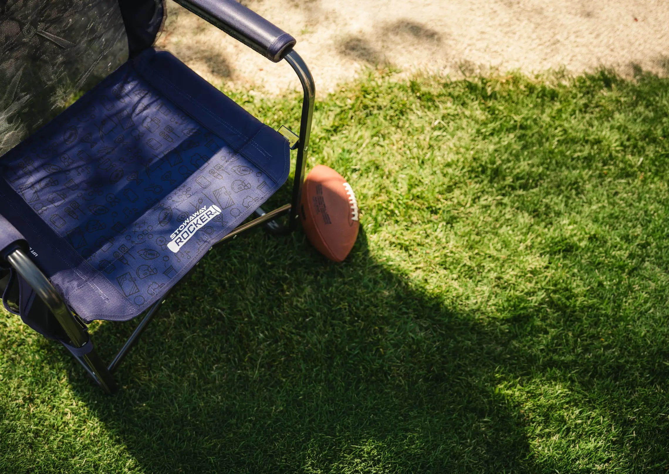 A tailgater stowaway rocker chair with a patterned seat cushion sits on green grass next to a brown football, with a tree trunk and dappled sunlight in the background.