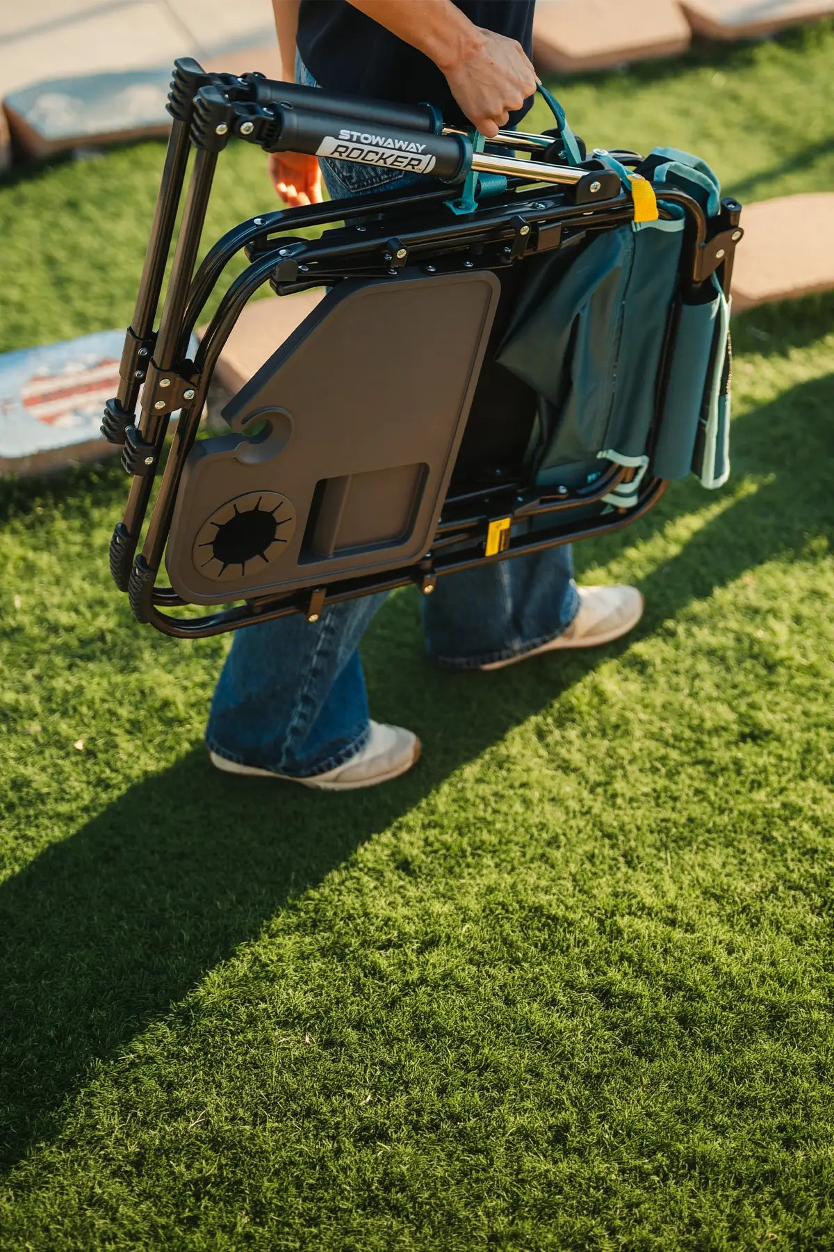 A person carrying the teal Stowaway Rocker with Side Table across a gras turf. 