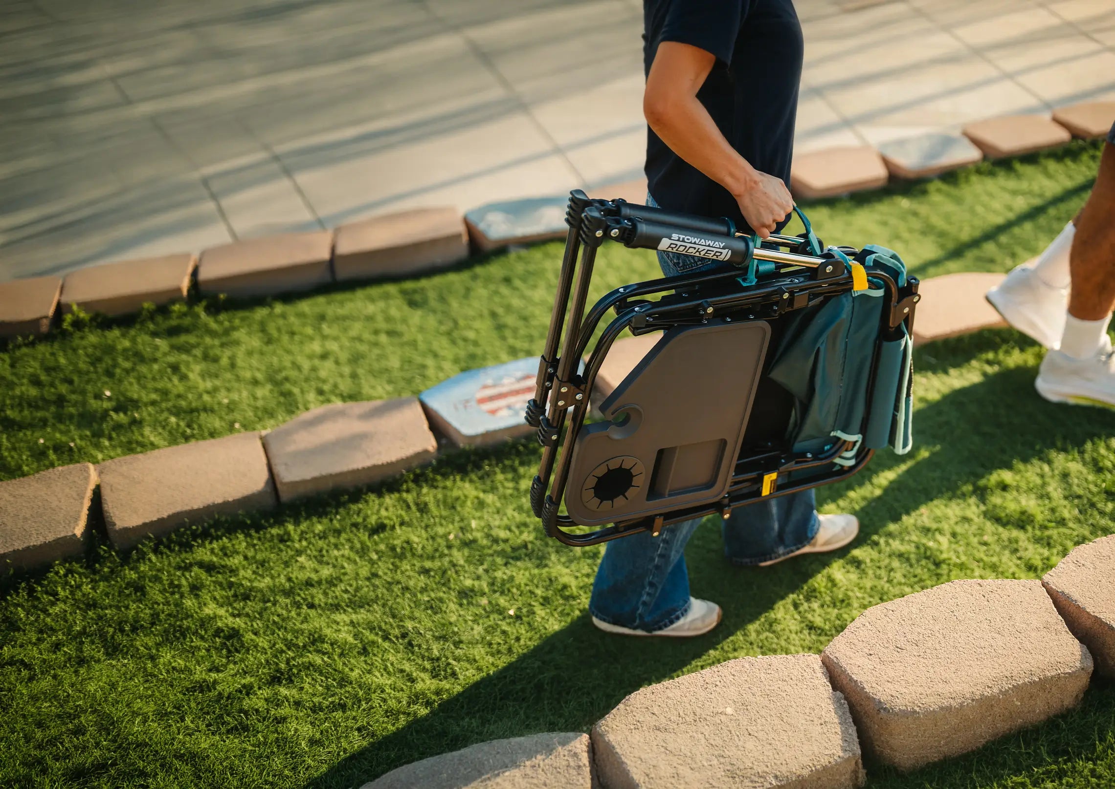 A person carrying the teal Stowaway Rocker with Side Table across a gras turf. 