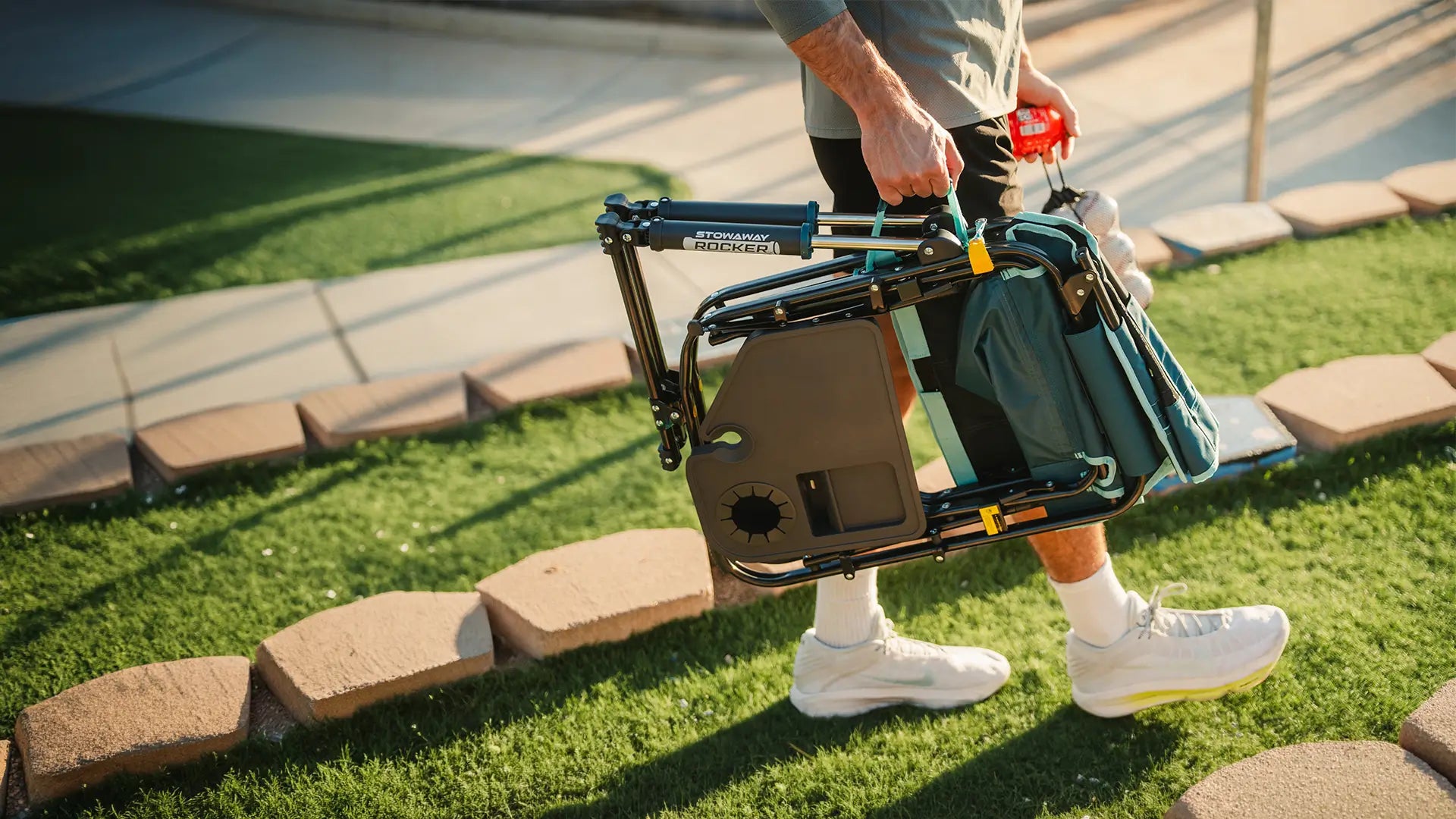 A man carrying a teal Stowaway Rocker with Side Table across a patch of grass. 